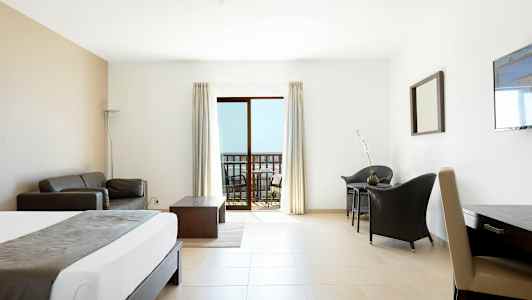 Ett hotellrum med ljus inredning. En säng, soffa, bord och stolar. En balkong med utsikt visas genom glasdörrar. (AI-genererad text)