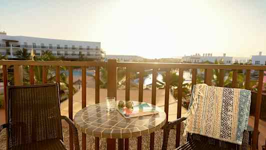 En balkong med två stolar, ett bord och utsikt över en pool och hotellbyggnader. (AI-genererad text)