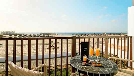 En balkong med utsikt över en strand och havet, med två stolar och ett bord dekorerat med drycker och snacks. (AI-genererad text)