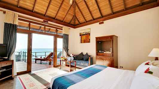 Ett hotellrum med en stor säng, ett träbord och en soffa, havsutsikt genom glasdörrar. (AI-genererad text)