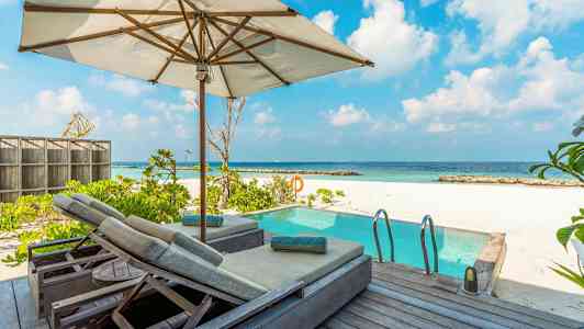 Bild av en soldäck med parasoll och solstolar bredvid en pool, med vit sandstrand och hav i bakgrunden. (AI-genererad text)