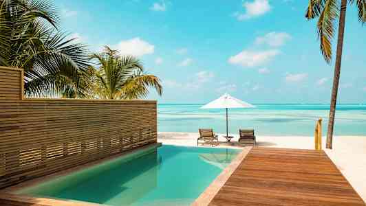 En pool bredvid en träterrass med utsikt över en strand med parasoll och solstolar. (AI-genererad text)