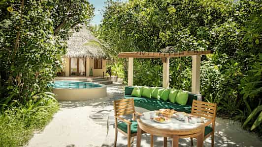 En trädgård med grönskande växter, ett runt poolområde, ett halmtäckt hus, och en sittgrupp med bord och stolar. (AI-genererad text)