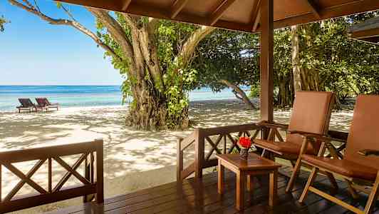 Utsikt från en veranda med stolar och bord mot en strand med solstolar och hav. (AI-genererad text)