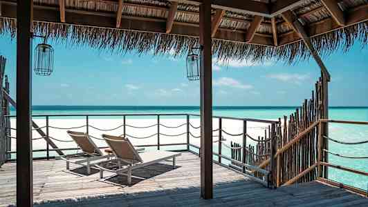 Två solstolar på en träbrygga med utsikt över en klarblå havskust under ett halmtak. (AI-genererad text)