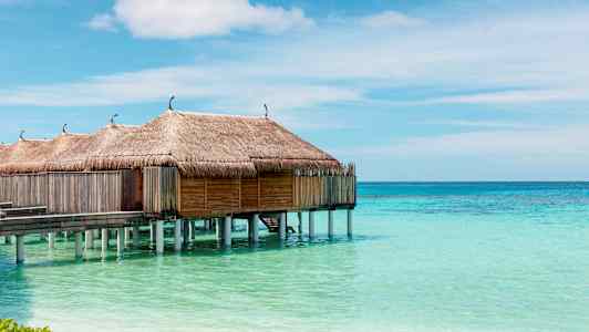 Vattenbungalows med halmtak på pålar över turkost vatten och klar himmel. (AI-genererad text)