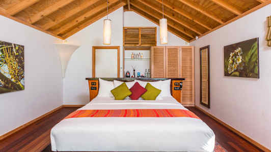 Ett sovrum med trätak, en stor säng med vita lakan och färgglada dekorativa kuddar, samt inredning inklusive lampor och väggdekorationer. (AI-genererad text)