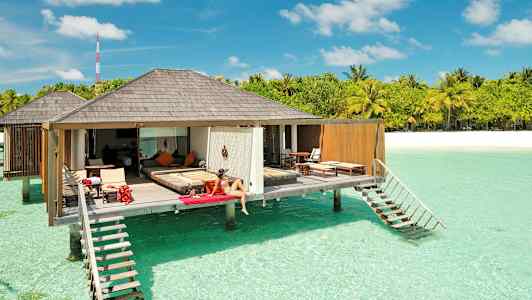 En kvinna sitter på en vilstol vid en lyxig bungalow som är placerad över klart turkost vatten, med tropiska träd och en sandstrand i bakgrunden. (AI-genererad text)