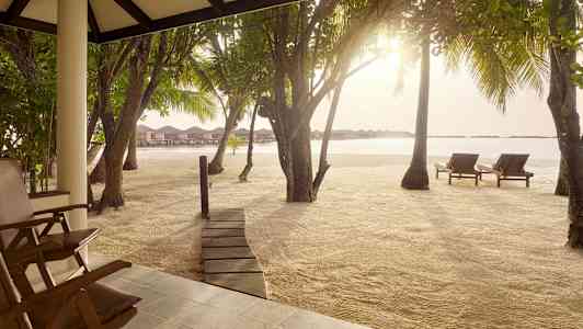 Vy från en terrass mot en sandstrand med solstolar, träd och bungalows på vattnet i bakgrunden. (AI-genererad text)