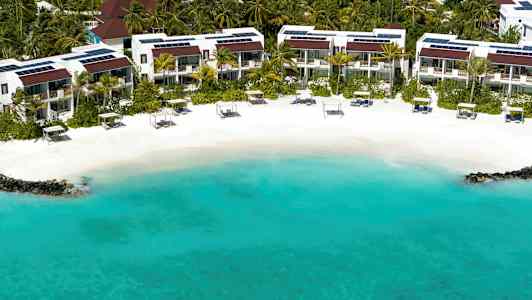 En strand med turkost vatten, vit sand och solstolar framför ett bostadsområde omgiven av palmer. (AI-genererad text)