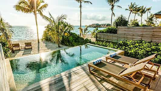 En pool med solstolar på en terrass vid en sandstrand och havet. (AI-genererad text)