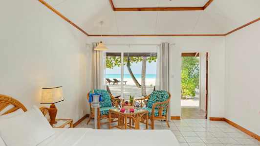 Ett hotellrum med säng, två stolar och bord visar utsikt mot en strand genom glasdörrar. (AI-genererad text)