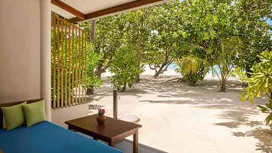 Vy från en terrass med en blå vilsoffa och ett bord med en vas, omgiven av sand och grönskande träd bredvid havet. (AI-genererad text)