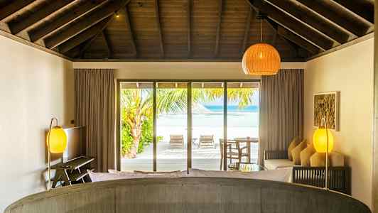 Rum med takbjälkar och utsikt över stranden genom stora glasdörrar. (AI-genererad text)