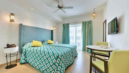 Ett hotellrum med två enkelsängar, en liten matplats och en vägg-TV framför fönster med färgglada gardiner. (AI-genererad text)