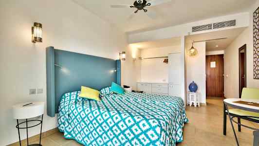 Ett hotellrum med en dubbelsäng, blått huvudgavel, färgglada kuddar och ett mönstrat täcke. Detaljer inkluderar möbler och dekorativa element. (AI-genererad text)