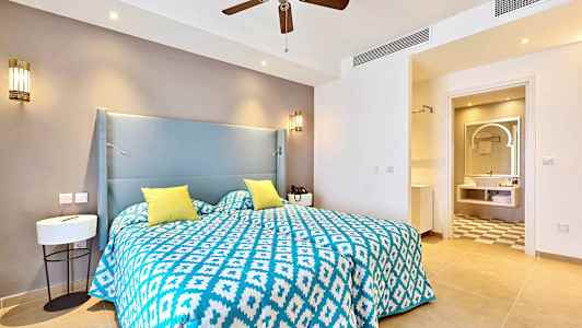 Ett hotellrum med en säng som har ett blått och vitt mönstrat överkast samt gula kuddar. I bakgrunden syns ett badrum. (AI-genererad text)