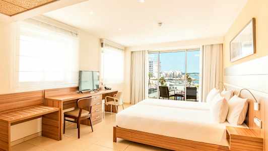 Ett ljust hotellrum med en stor säng, arbetsbord och tv, samt en balkong med utsikt över en stad och havet. (AI-genererad text)