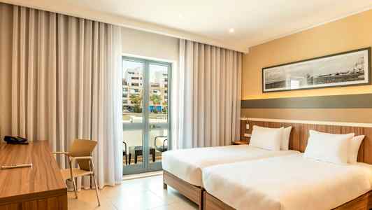 Hotellrum med två enkelsängar, skrivbord och en balkong med glasdörrar. (AI-genererad text)