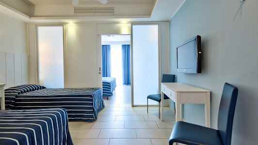 Två enkelbäddar med randiga blå överkast, ett vitt skrivbord med två blå stolar och en väggmonterad TV, samt en glaserad dörr till rummet bakom. (AI-genererad text)