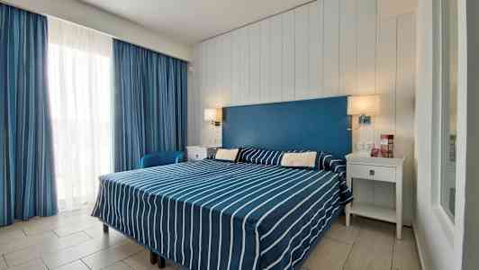 Ett hotellrum med en dubbelsäng klädd i blå- och vitrandiga sängkläder, en blå sänggavel och vita möbler. (AI-genererad text)