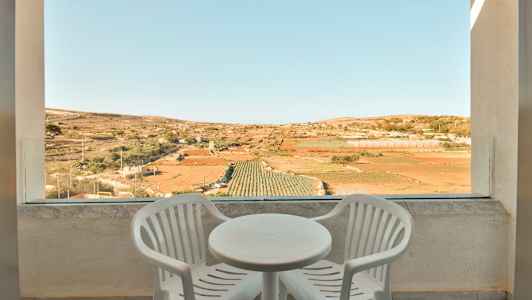 Två vita plaststolar och ett runt bord på en terrass med utsikt över odlingsfält. (AI-genererad text)