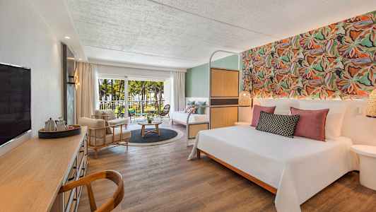 Hotellrum med dubbelsäng, sittgrupp, skrivbord och balkong med utsikt över palmer och trädgård. (AI-genererad text)