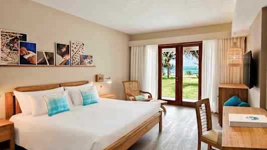 Ett hotellrum med en dubbelsäng, träinredning, och en glasdörr som öppnar upp till en grön trädgård och havsutsikt. (AI-genererad text)