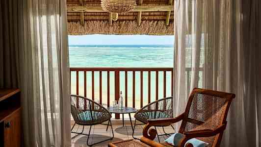 Utsikt över havet från ett rum med en taktäckt balkong. Mellan transparenta gardiner syns två trädgårdsstolar vid ett bord. (AI-genererad text)