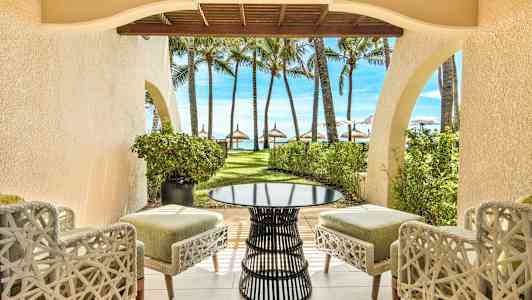 Utsikt från en uteplats med stolar och ett bord mot en trädgård med palmer och strand i bakgrunden. (AI-genererad text)