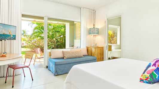 Ett ljust hotellrum med säng, sittgrupp och bord. Utsikt mot trädgården syns genom glasdörrarna. (AI-genererad text)