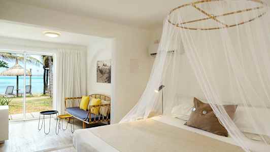 Hotellrum med en stor säng täckt med ett myggnät, en soffa, bord och ett stort fönster med utsikt över stranden och havet. (AI-genererad text)