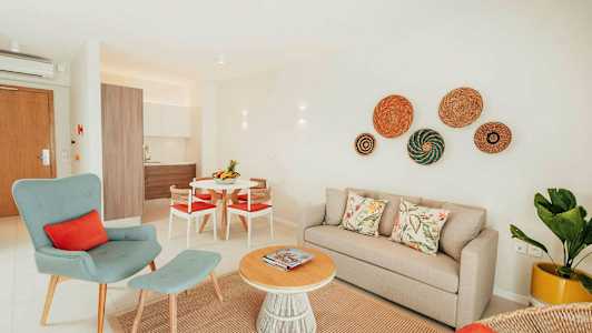 Ett ljust och modernt inrett vardagsrum med beige soffa, grön fåtölj och ett runt soffbord av trä. Köksdelen syns i bakgrunden. (AI-genererad text)