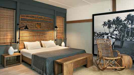 Ett hotellrum med en dubbelsäng, trädetaljer och en stol av rotting. En stor tavla med svartvitt motiv pryder väggen. (AI-genererad text)