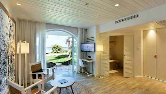 Ett hotellrum med sittmöbler, ett bord och en TV. En dörr leder till ett badrum, och det finns fönster som vetter mot en grön trädgård och stranden. (AI-genererad text)