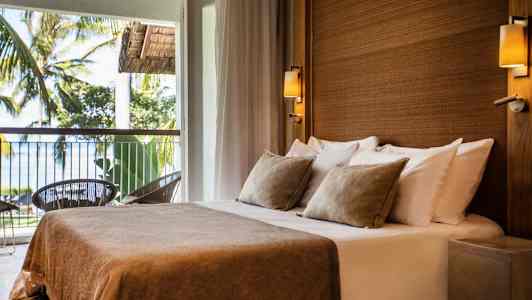 Ett rum med en dubbelsäng som har beige sängkläder och kuddar, beläget vid en balkong med utsikt över träd och hav. (AI-genererad text)