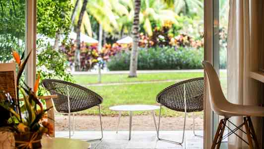 Utsikt från ett rum genom en öppen dörr mot en uteplats med två stolar och ett bord, omgiven av grönområde och palmer. (AI-genererad text)
