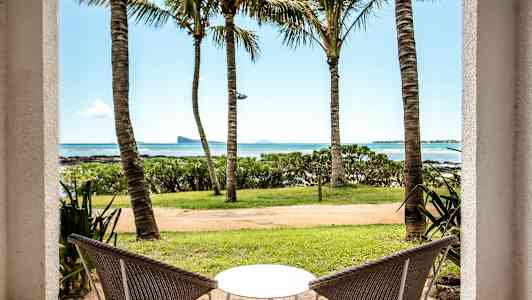 Två stolar och ett bord med utsikt över havet genom palmer. (AI-genererad text)