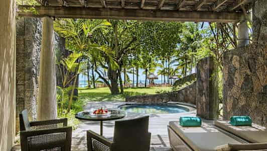 En terrass med sittplatser och solstolar bredvid en liten pool, omgiven av växter och med utsikt mot stranden och palmträd. (AI-genererad text)