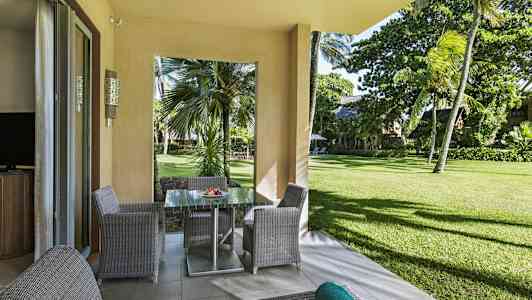 Utsikt från en terrass med trädgårdsmöbler mot grönska och palmer. (AI-genererad text)