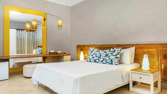 Ett hotellrum med en säng försedd med dekorativa kuddar, ett nattbord med lampa och en stor spegel med gul ram. (AI-genererad text)