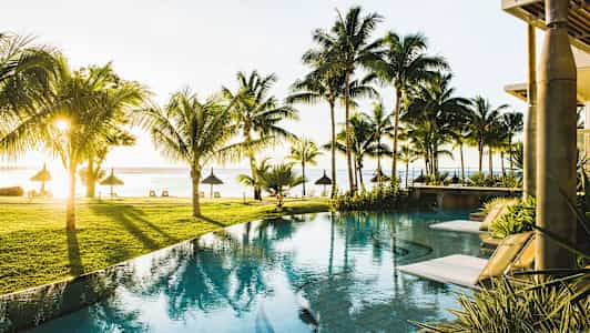 Vy över en pool med solstolar kantad av palmer och utsikt över stranden vid havet. (AI-genererad text)