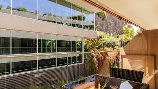 Uteplats med möbler framför modernt glasbyggnad vid en grönskande bergvägg. (AI-genererad text)