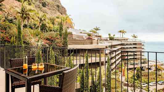 Bild av en balkong med ett möblemang av rotting, med ett bord som har två glas och en karaff. Utsikt mot en plantering, byggnader och havet. (AI-genererad text)