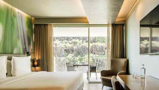 Ett hotellrum med en dubbelsäng, sittgrupp, ett bord med stolar samt en stor glasdörr som öppnar upp till en balkong med utsikt över natur och klippor. (AI-genererad text)