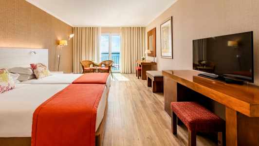 Ett hotellrum med två sängar, träbord och en TV, gardiner och en balkong med utsikt mot havet. (AI-genererad text)