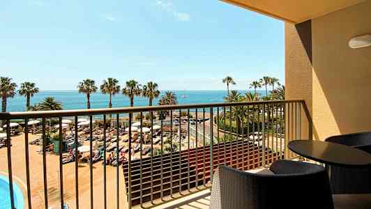 Utsikt från en balkong med möbler över ett poolområde, palmer och havet i bakgrunden. (AI-genererad text)
