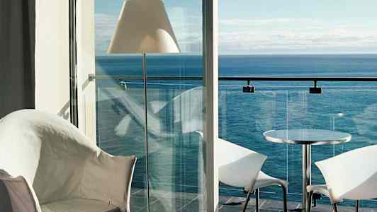 Utsikt från ett rum mot havet, med en balkong med glasräcke, bord och stolar. (AI-genererad text)