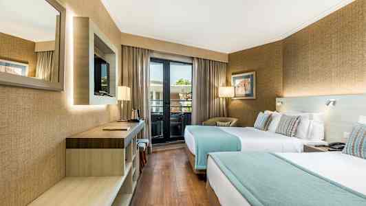 Ett hotellrum med två enkelsängar, en skrivbordsyta, en väggmonterad TV och ett fönster som leder till en balkong. (AI-genererad text)