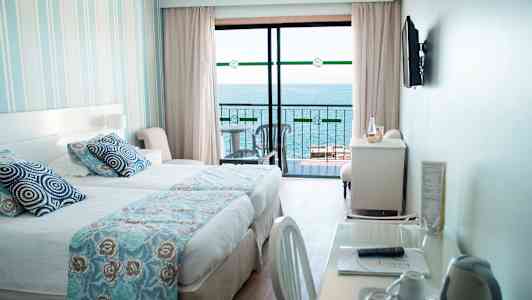 Ett hotellrum med en säng, ett skrivbord och utsikt över havet från en balkong. (AI-genererad text)